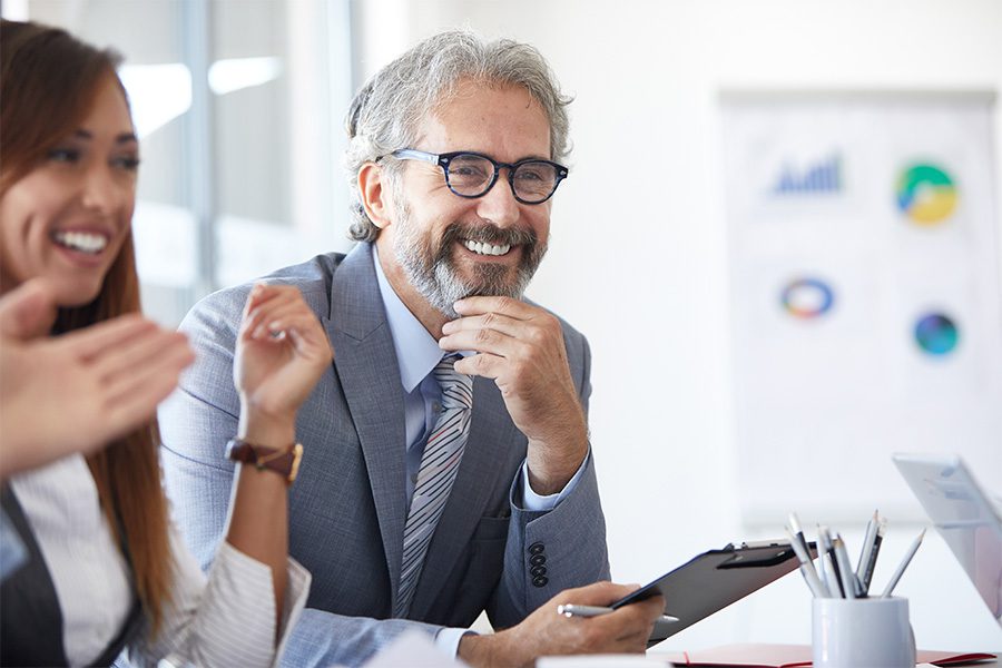 Agency Partnerships - Business Owner Smiling While Holding a Clipboard and Listening During a Team Meeting